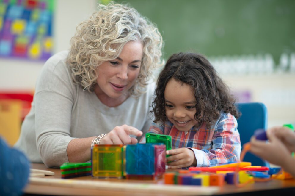 A boy of African descent sits at a table in his classroom one afternoon. Him and his teacher are building a cube together out of magnetic plastic building pieces. They are focused but also smiling and having fun.