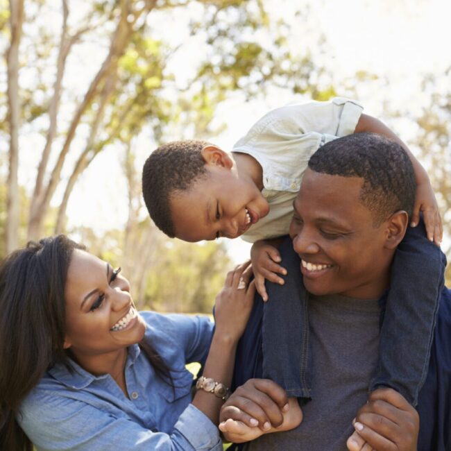 Parents Carrying Son On Shoulders As They Walk In Park
